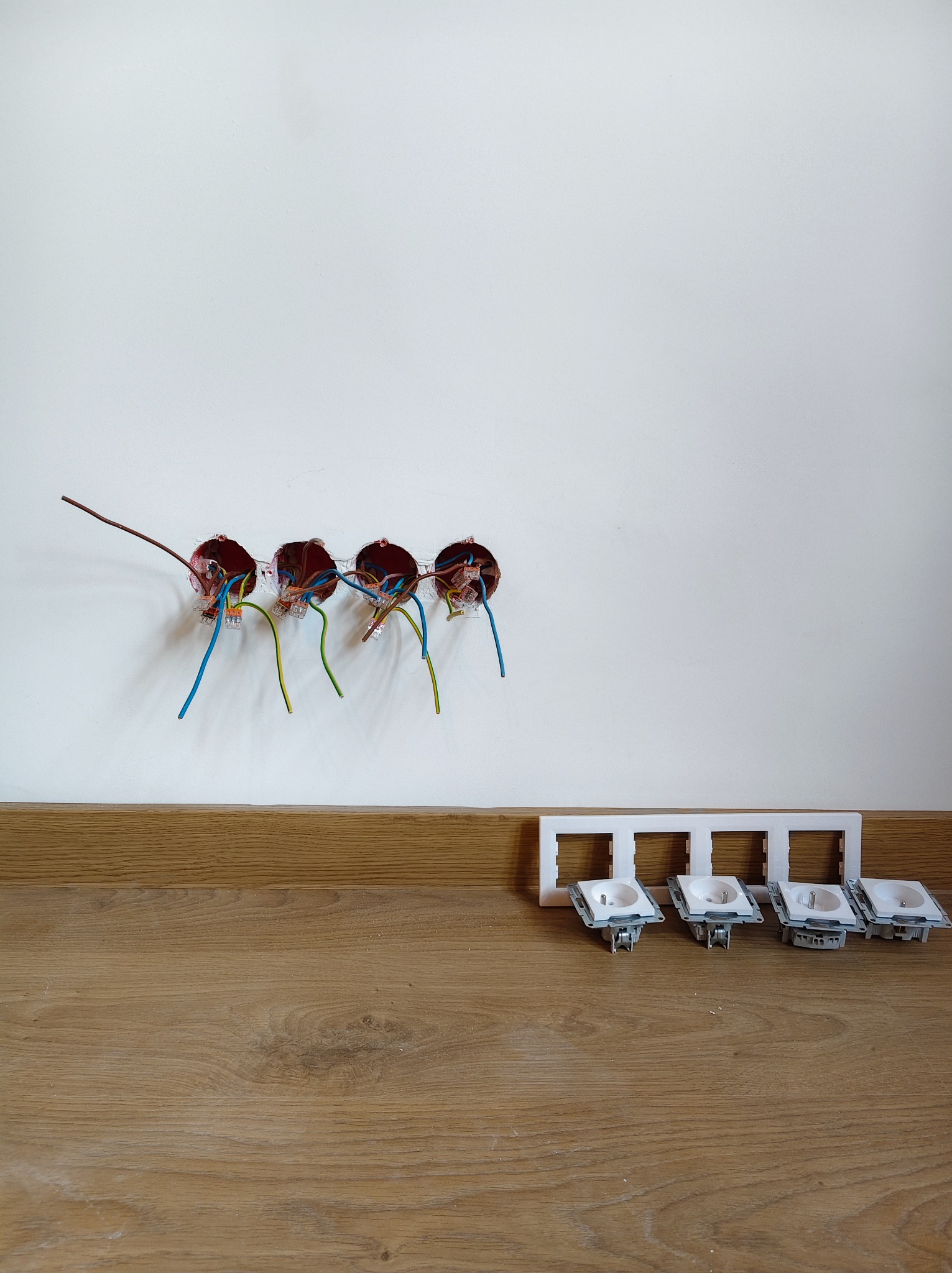 Cztery puszki elektryczne w ścianie z podłączonymi przewodami, obok leżą gniazdka z ramką, gotowe do montażu. Widok na drewnianą podłogę i białą ścianę.