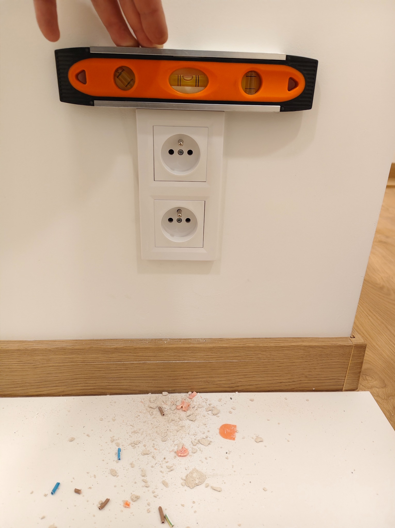 Poziomica przykładana do gniazdka elektrycznego podwójnego, białe gniazdko na białej ścianie, widoczne ślady po montażu.