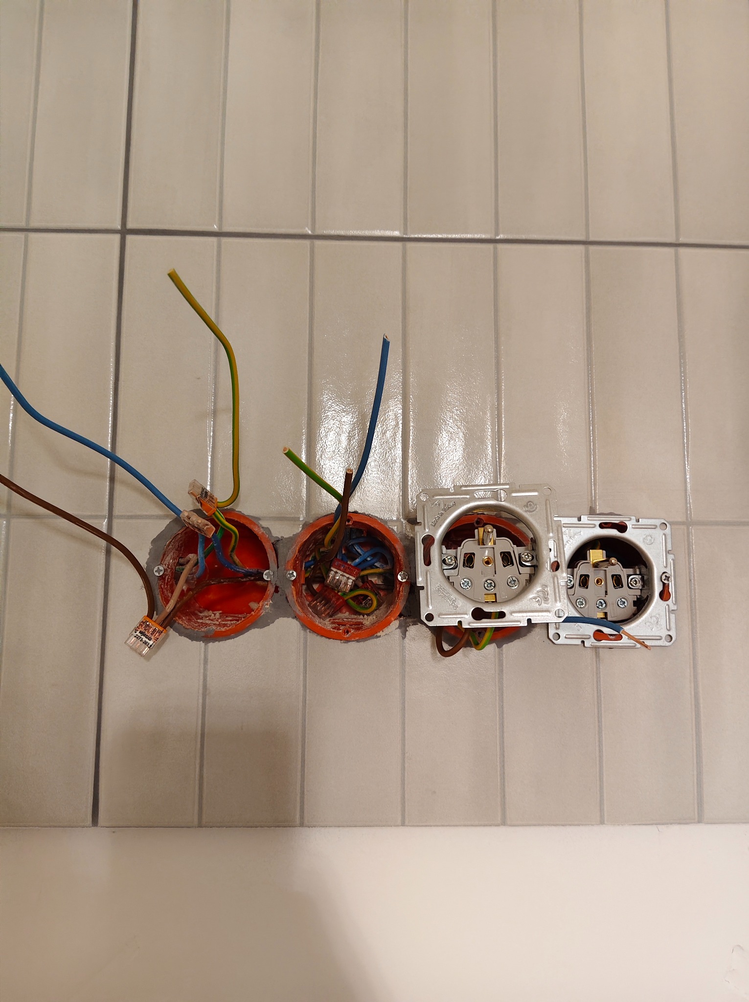 Otwory w ścianie z wystającymi przewodami i zamontowanymi puszkami elektrycznymi, widoczne złącza WAGO. Prace instalacyjne na tle płytek ceramicznych.