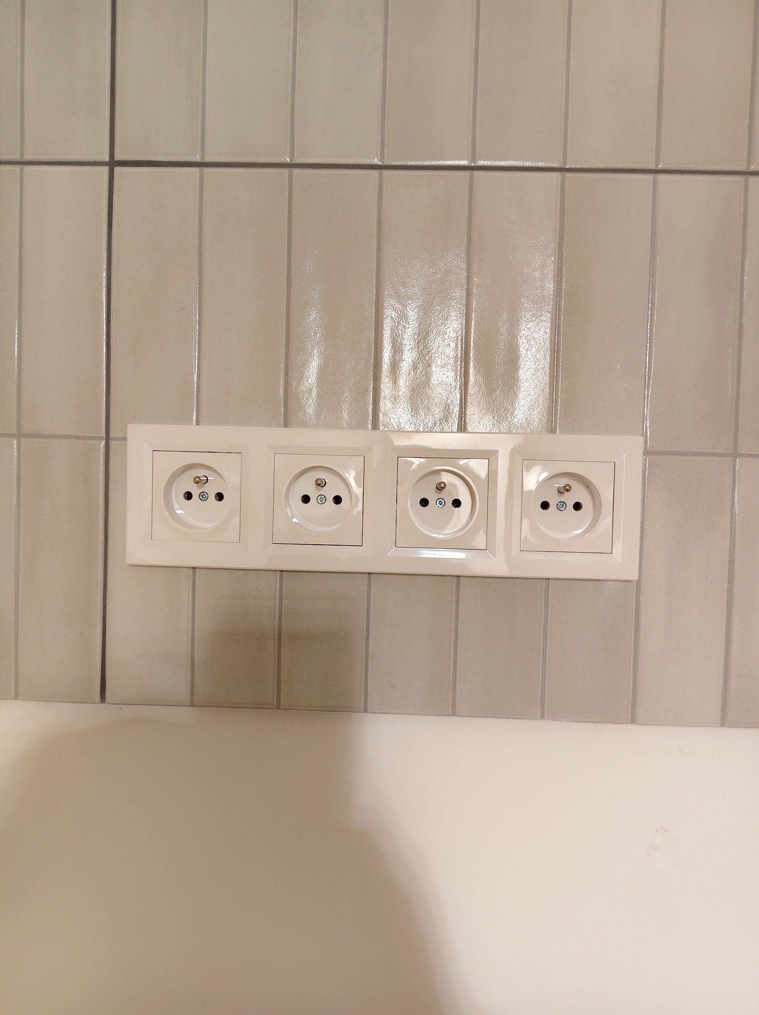 Cztery gniazdka elektryczne w białej ramce, zamontowane na ścianie wyłożonej błyszczącymi, pionowymi płytkami w odcieniach szarości. Widoczny fragment białej ściany poniżej.