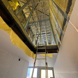 Sławomir Wojtuń - Skośny kadr na poddasze w trakcie adaptacji: widoczne ocieplenie wełną mineralną, metalowa konstrukcja szkieletowa, częściowo obłożona płytami gipsowo-kartonowymi i drabina.