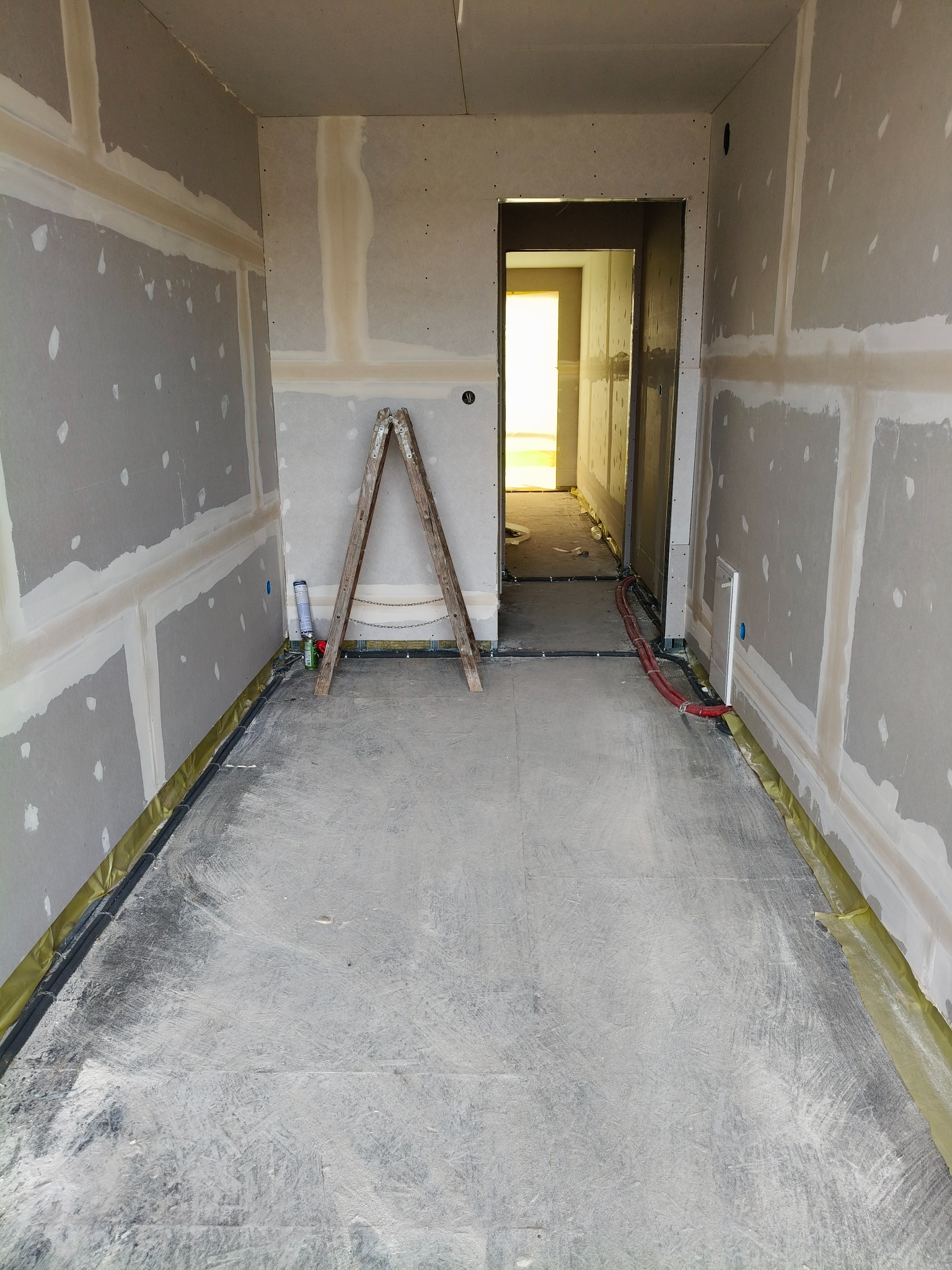 Wnętrze w trakcie prac wykończeniowych: ściany z płyt kartonowo-gipsowych, widoczne łączenia, drabina, instalacje podłogowe. Surowy stan przed dalszymi pracami.