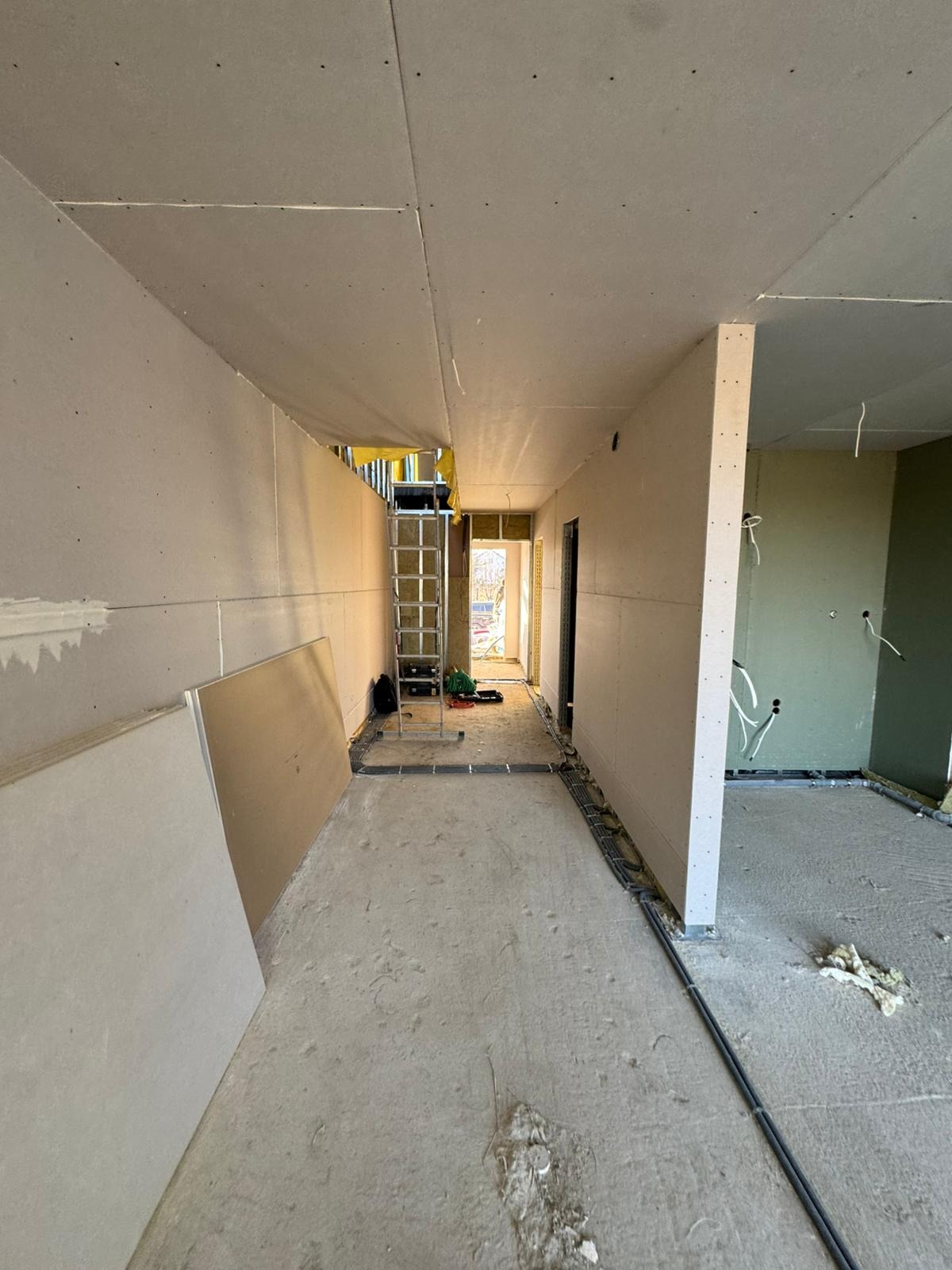 Korytarz w trakcie montażu płyt gipsowo-kartonowych na ścianach i suficie. Widoczna drabina, narzędzia i instalacje elektryczne. Surowy stan wykończenia.