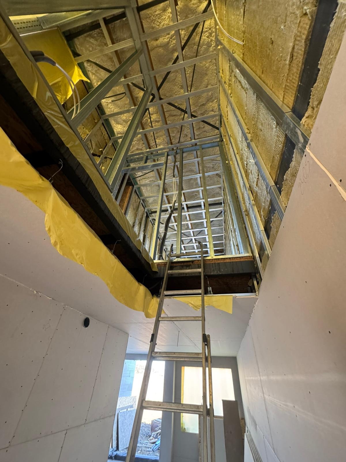 Skośny kadr na poddasze w trakcie adaptacji: widoczne ocieplenie wełną mineralną, metalowa konstrukcja szkieletowa, częściowo obłożona płytami gipsowo-kartonowymi i drabina.