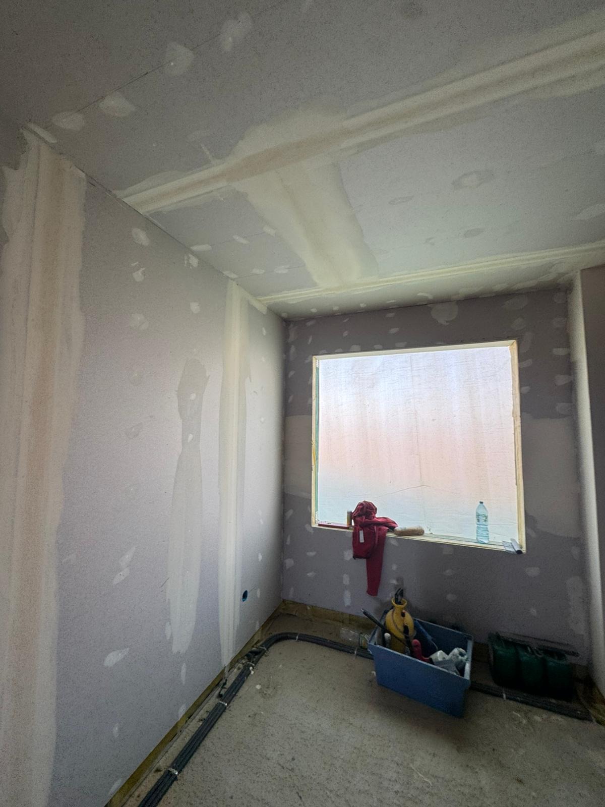 Pokój w trakcie prac wykończeniowych: ściany przygotowane do malowania, widoczne łączenia płyt, narzędzia i instalacja elektryczna przy podłodze.