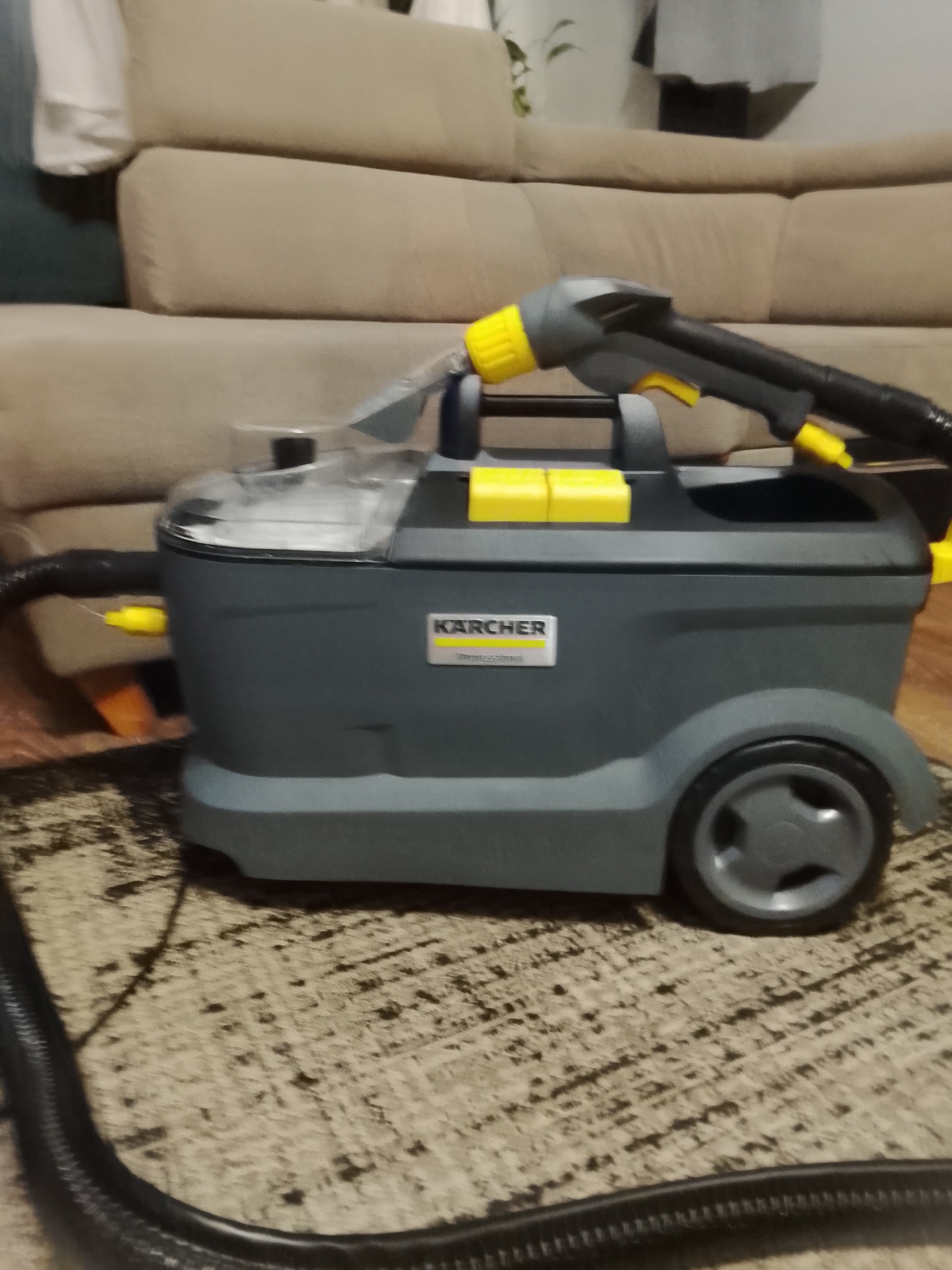 Szaro-żółty odkurzacz piorący Karcher na tle beżowej kanapy i wzorzystego dywanu. Widoczny wąż i dysza urządzenia. Sprzęt gotowy do użycia.