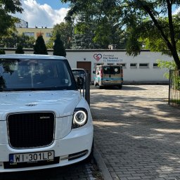 Podróż Bez Barier - Biała taksówka elektryczna LEVC TX z warszawską rejestracją WI 301LP na tle budynku Fundacji Daj Szansę. W tle zaparkowany bus.