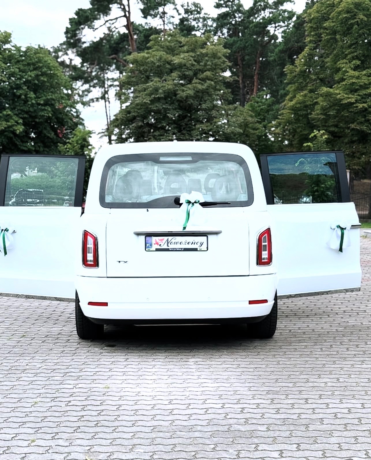 Biały, luksusowy samochód z otwartymi drzwiami, udekorowany zielonymi wstążkami, tablica rejestracyjna z napisem 'Nowożeńcy', tło: zieleń drzew.