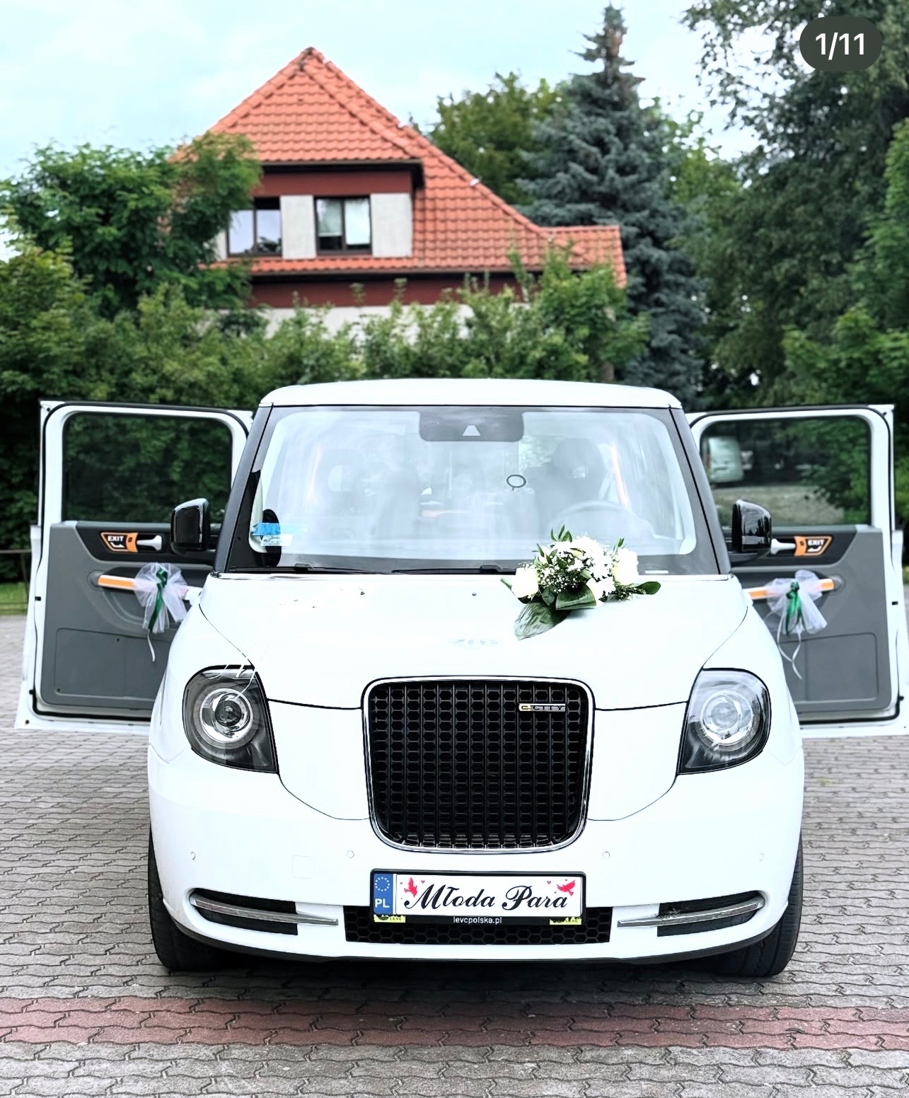 Biały, luksusowy samochód z otwartymi drzwiami, udekorowany kwiatami, zaparkowany na brukowanej nawierzchni. W tle dom z czerwoną dachówką i zieleń.