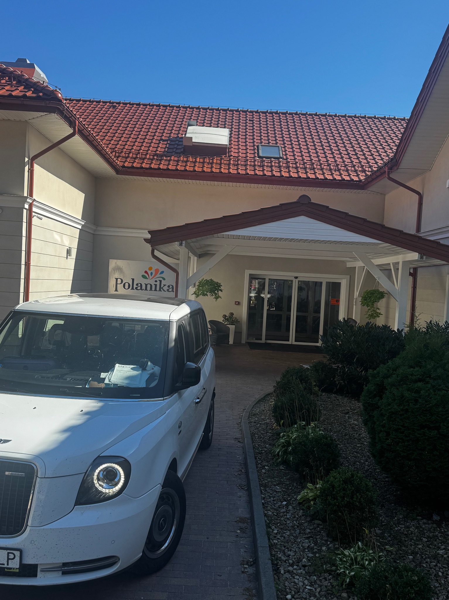 Biała taksówka TX4 przed budynkiem z szyldem 'Polanika' w słoneczny dzień. Charakterystyczny design pojazdu.