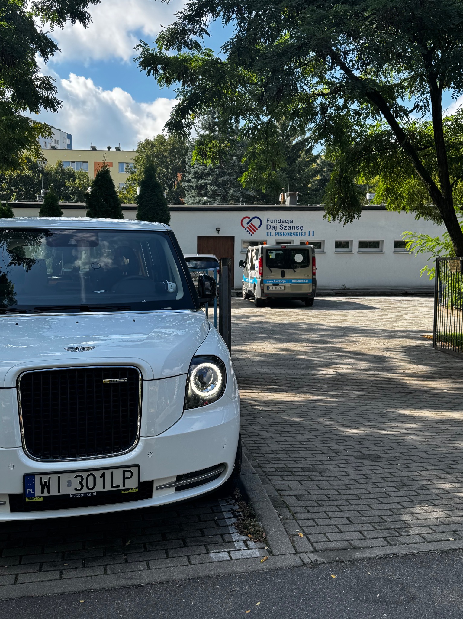 Biała taksówka elektryczna LEVC TX z warszawską rejestracją WI 301LP na tle budynku Fundacji Daj Szansę. W tle zaparkowany bus.