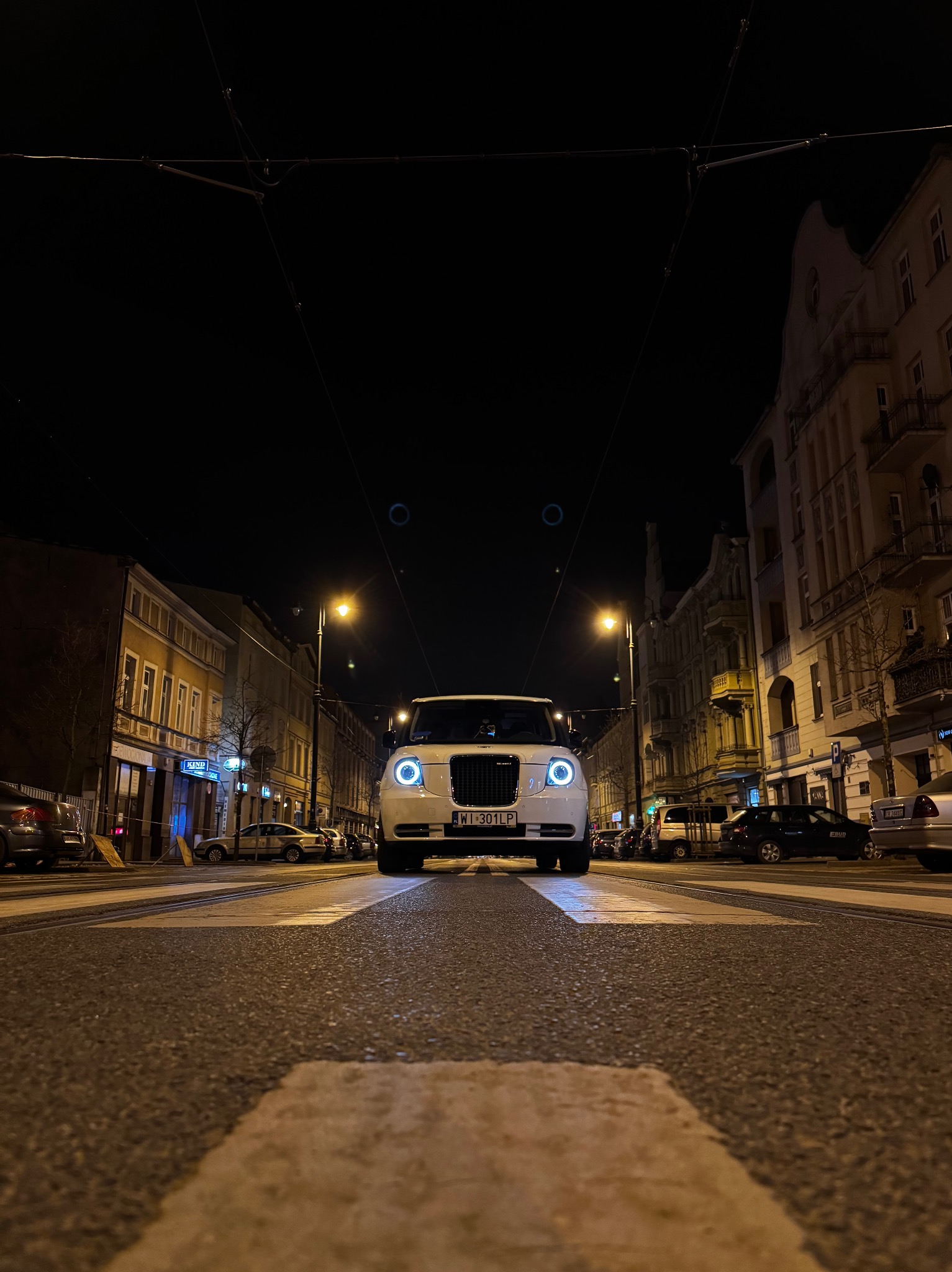 Biała taksówka w stylu londyńskim na pasach nocą, z charakterystycznymi okrągłymi światłami, widziana z niskiej perspektywy na tle miejskiej ulicy.