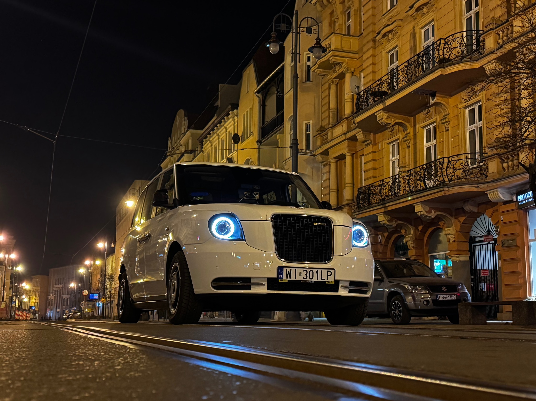 Biała taksówka w stylu retro z niebieskimi światłami LED na tle nocnej, oświetlonej ulicy z torami tramwajowymi i budynkami w tle.