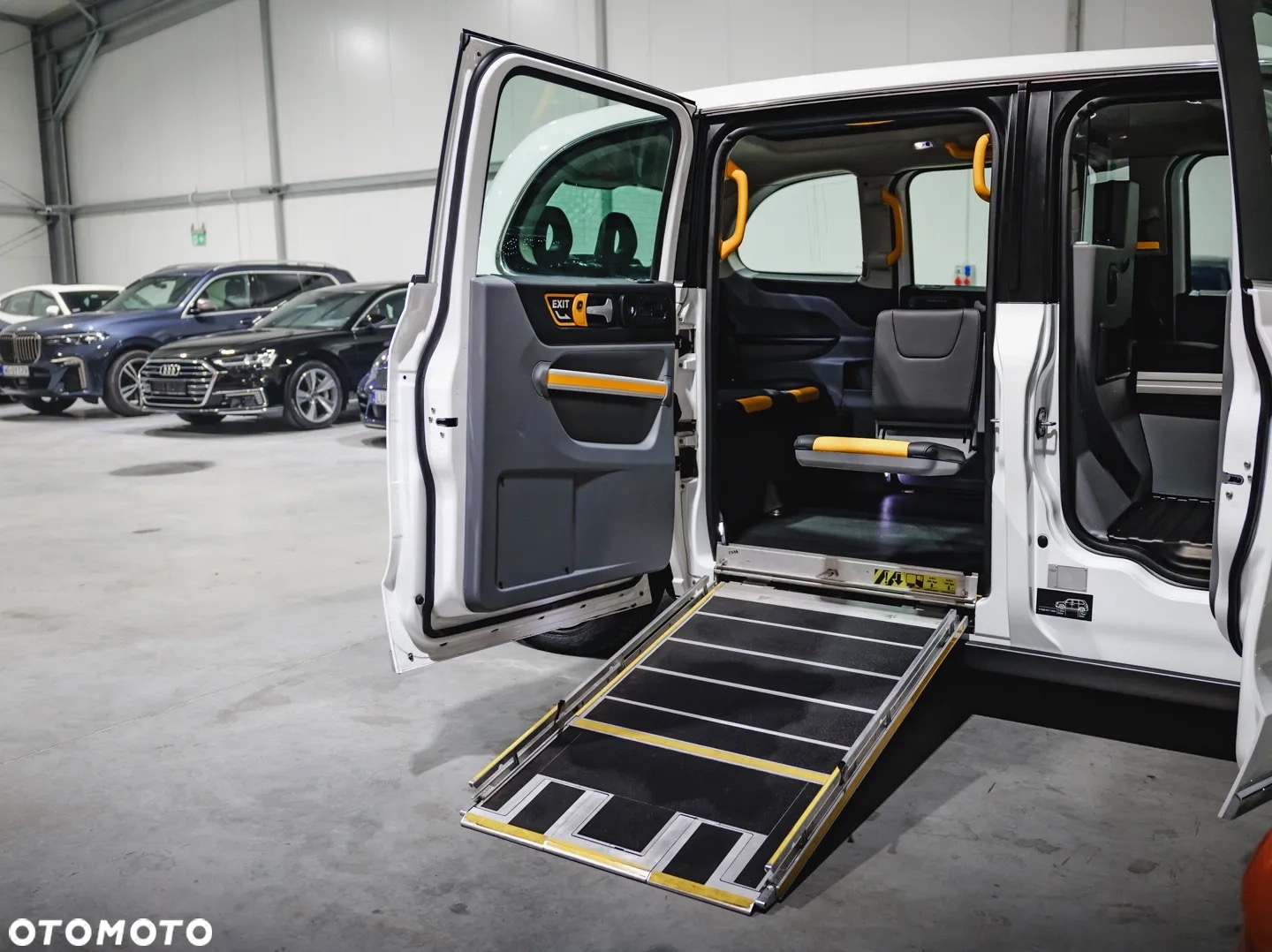 Wnętrze białego busa z rozłożoną rampą dla osób niepełnosprawnych. Widoczne fotele i uchwyty w kolorze żółtym. W tle zaparkowane samochody.