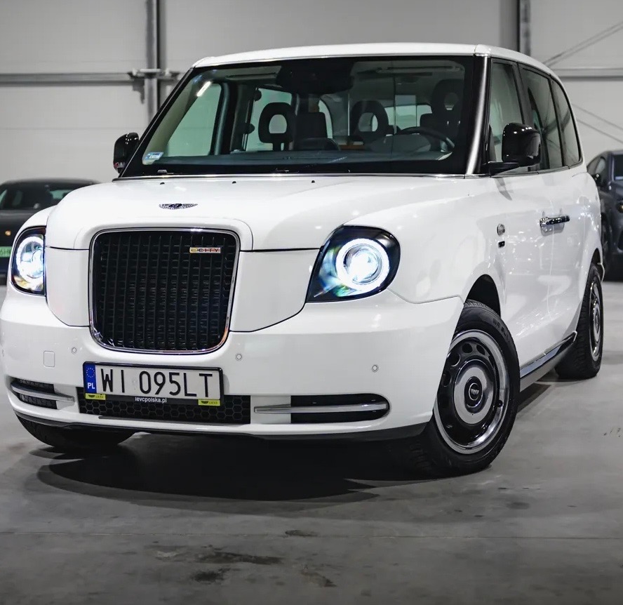 Biały, luksusowy bus z charakterystycznym grillem i okrągłymi reflektorami, zaparkowany w jasnym garażu. Widok z przodu, lekko z dołu, podkreślający nowoczesny design.