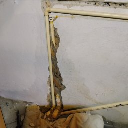 HYDROTOMI TOMASZ OSTAŁOWSKI - Narożne ułożenie beżowych rur PCV w ścianie z ubytkami tynku, widoczne narzędzia i fragment ręcznika. Instalacja po naprawie, z datą na dole zdjęcia.