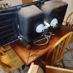 HYDROTOMI TOMASZ OSTAŁOWSKI - Czarny, dwukomorowy zlew kuchenny od spodu, z podłączonymi rurami i syfonem, leżący na drewnianym stole otoczonym krzesłami. Widoczne elementy instalacji.