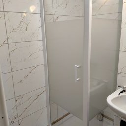 Hydrotomi - Nowoczesna kabina prysznicowa z matowego szkła i białymi profilami, umywalka z baterią w tle, płytki imitujące marmur na ścianach. Widok na łazienkę po remoncie.
