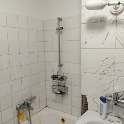 HYDROTOMI TOMASZ OSTAŁOWSKI - Zardzewiała bateria wannowa i słuchawka prysznicowa na tle białych, kwadratowych płytek. Widoczne ślady rdzy i niedoskonałości. Umywalka z brakującymi płytkami w tle.