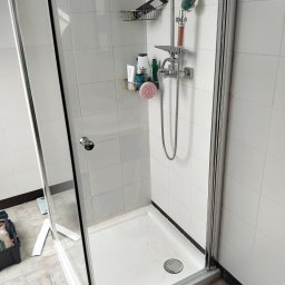 Hydrotomi - Kabina prysznicowa z chromowaną armaturą i półkami na kosmetyki na tle białych płytek. Widoczny brodzik i narzędzia hydrauliczne.