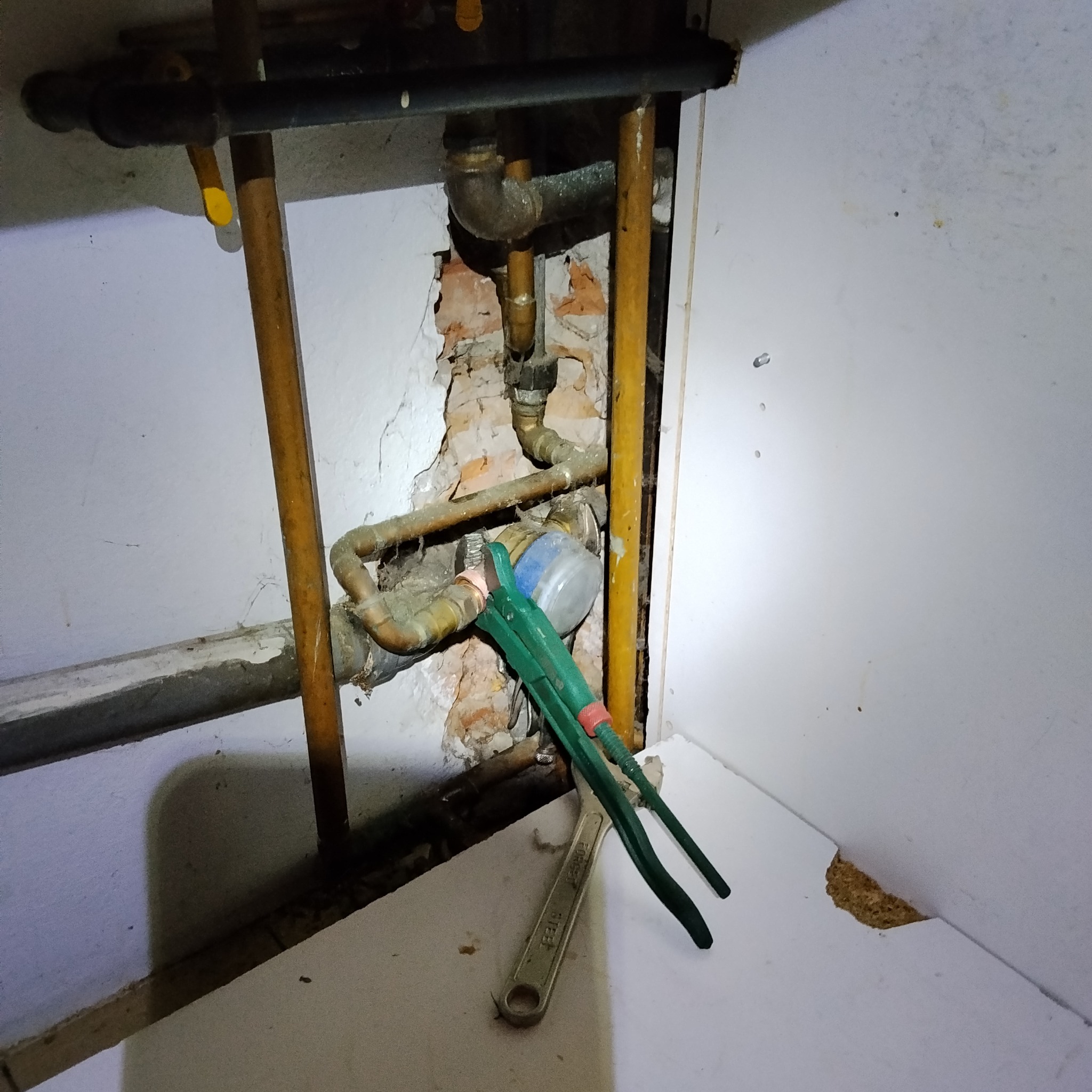 Odsłonięte rury miedziane w ścianie, z zielonym kluczem hydraulicznym i kluczem płaskim. Widoczne uszkodzenia tynku i cegły. Narożne ujęcie.