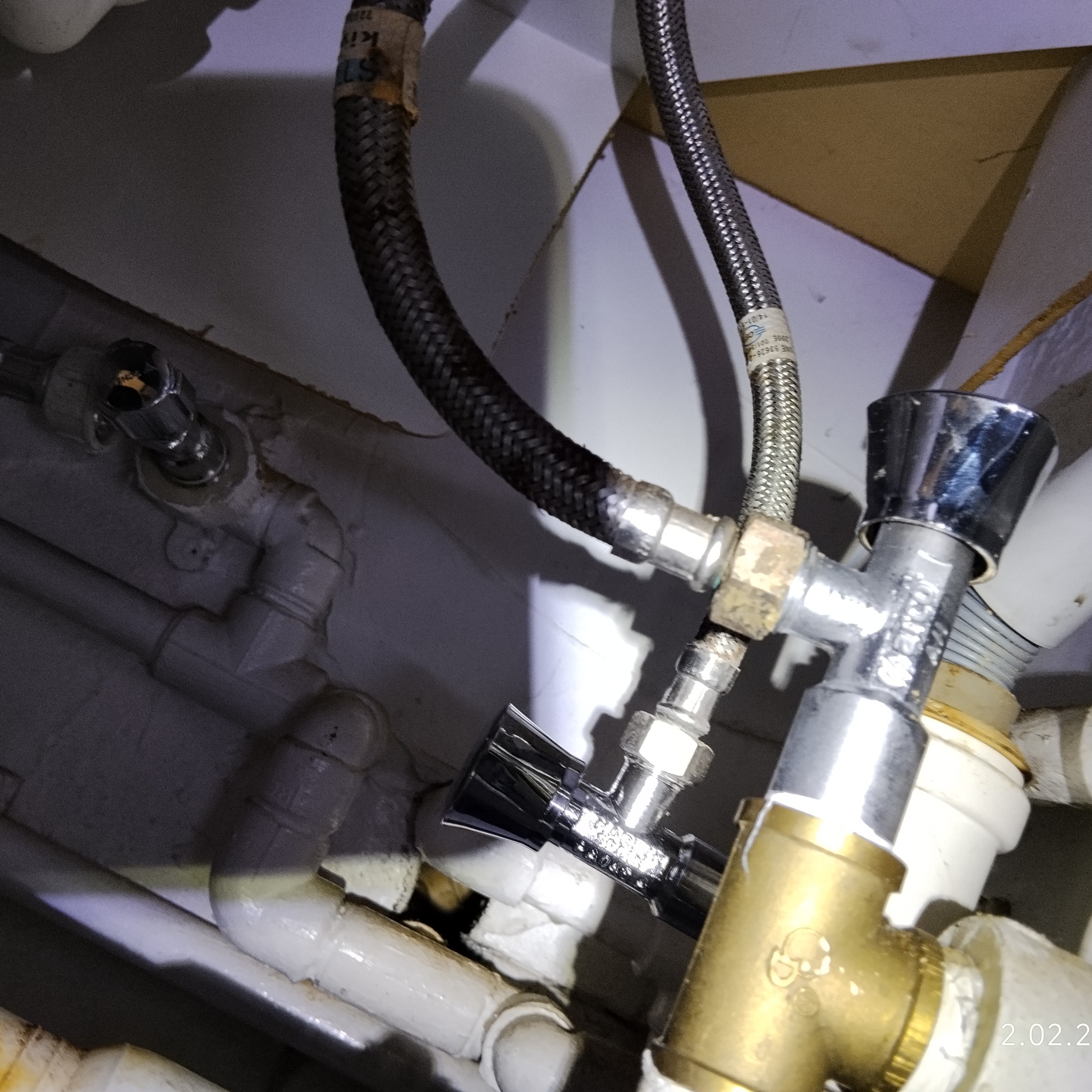 Błyszczący zawór kątowy z chromowaną rączką, zamontowany na białych rurach instalacji wodnej, z widocznymi elastycznymi wężami w oplocie.