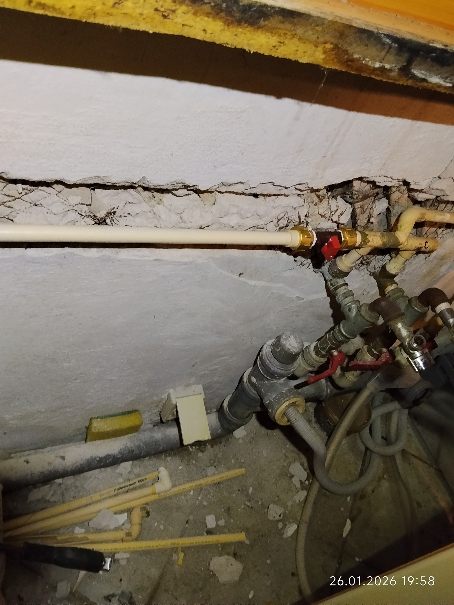 Złożony węzeł rur instalacji wodnej z zaworami, widoczny przyłącze do ściany z uszkodzonym tynkiem. Różne materiały i kolory rur. Na podłodze fragmenty gruzu.