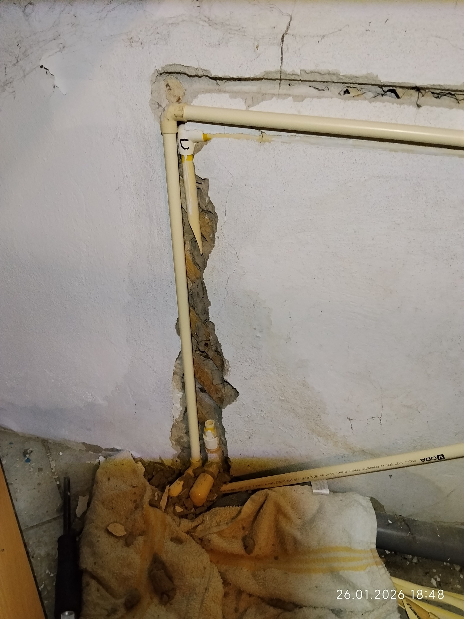 Narożne ułożenie beżowych rur PCV w ścianie z ubytkami tynku, widoczne narzędzia i fragment ręcznika. Instalacja po naprawie, z datą na dole zdjęcia.