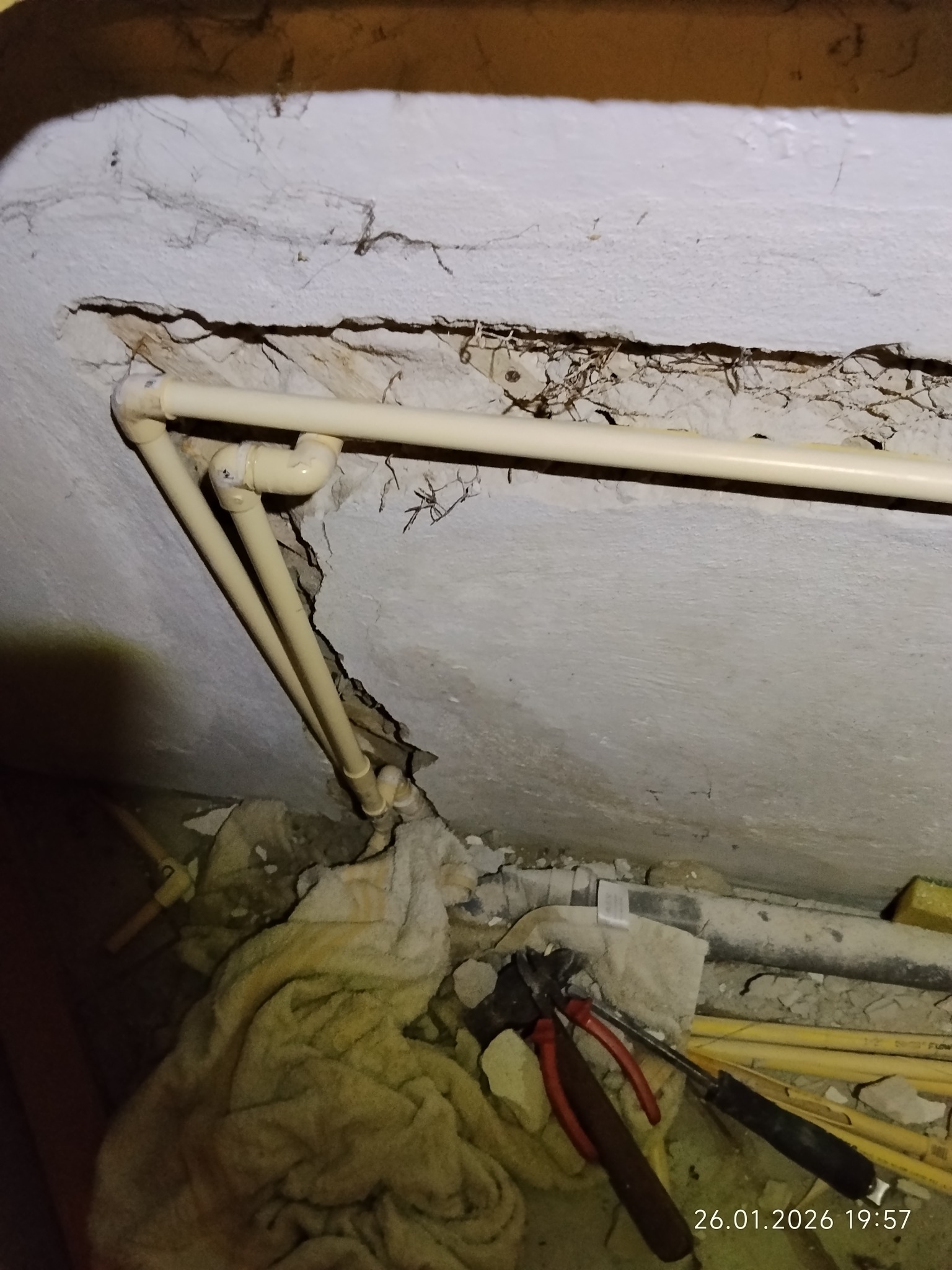 Narożne, beżowe rury instalacji wodnej, prowadzone wzdłuż ściany z ubytkiem tynku. Widoczne narzędzia hydrauliczne i fragment ręcznika. Ujęcie z dołu.