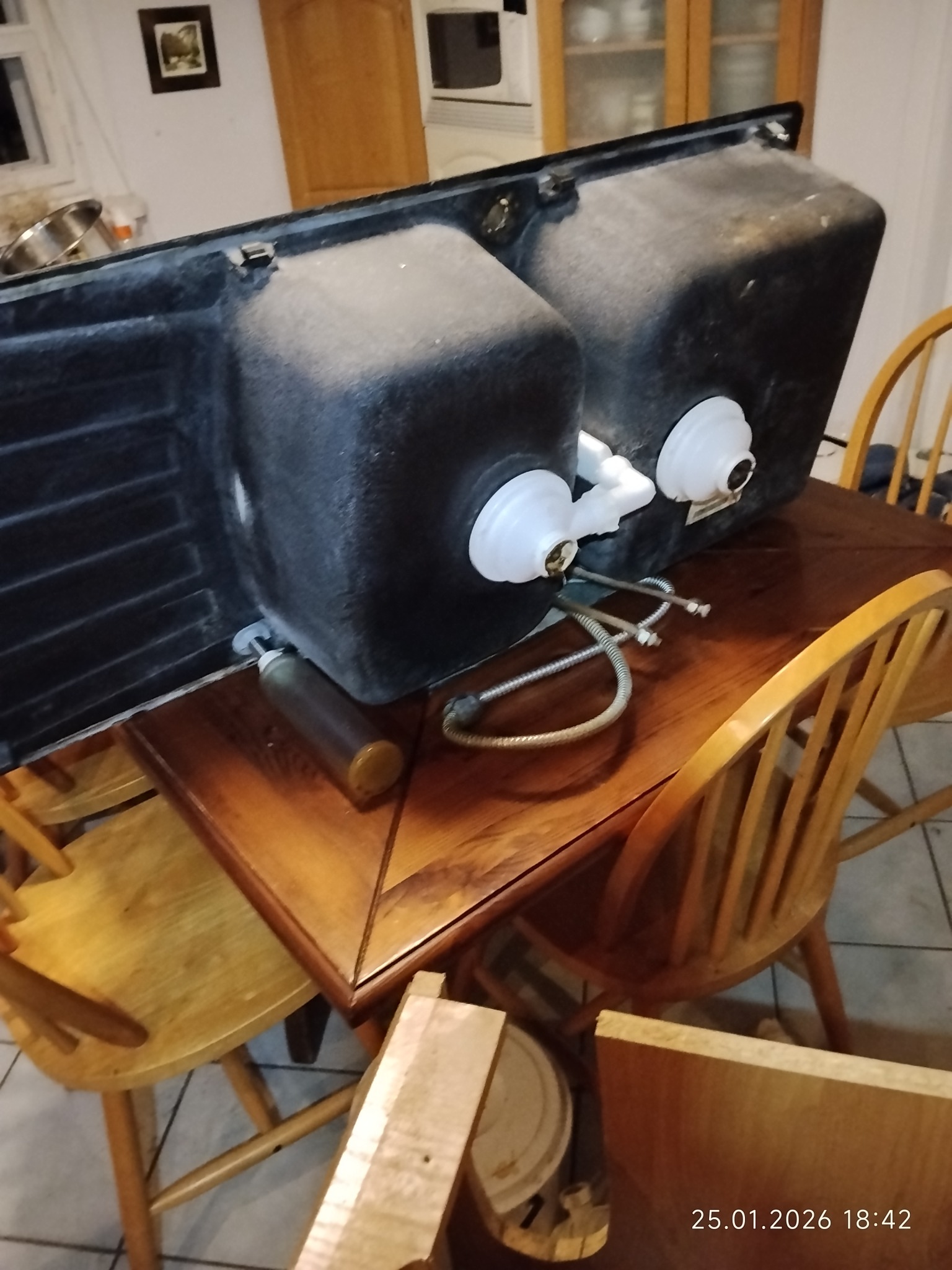 Czarny, dwukomorowy zlew kuchenny od spodu, z podłączonymi rurami i syfonem, leżący na drewnianym stole otoczonym krzesłami. Widoczne elementy instalacji.