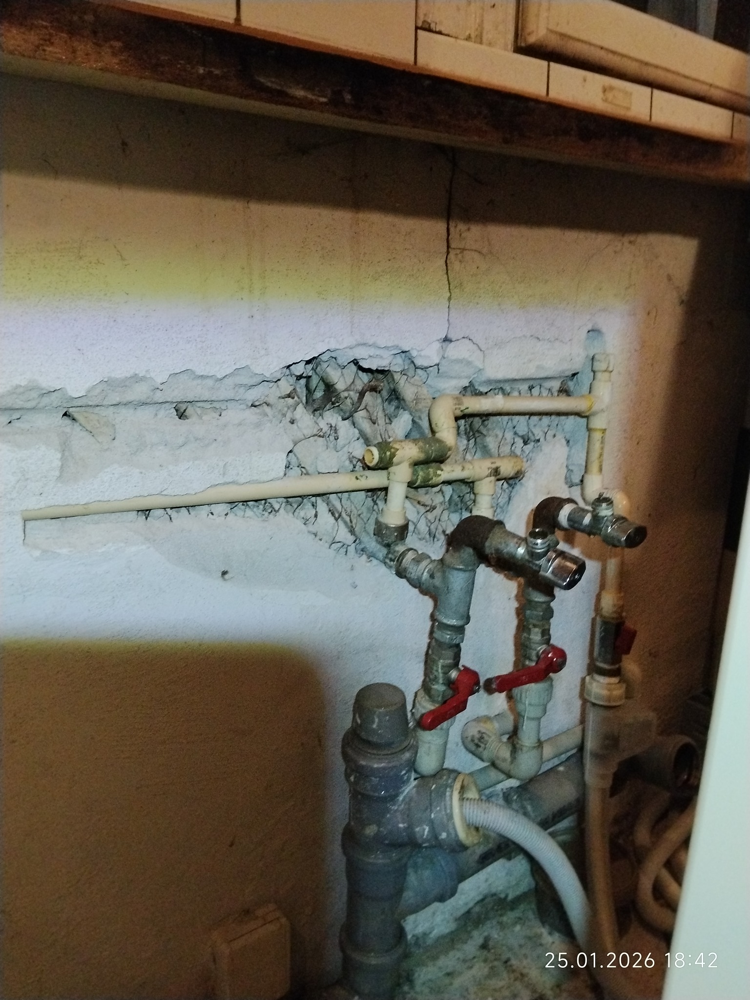 Fragment instalacji hydraulicznej z rurami w ścianie, widoczne zawory z czerwonymi uchwytami i uszkodzony tynk. Różne materiały rur i złączki.