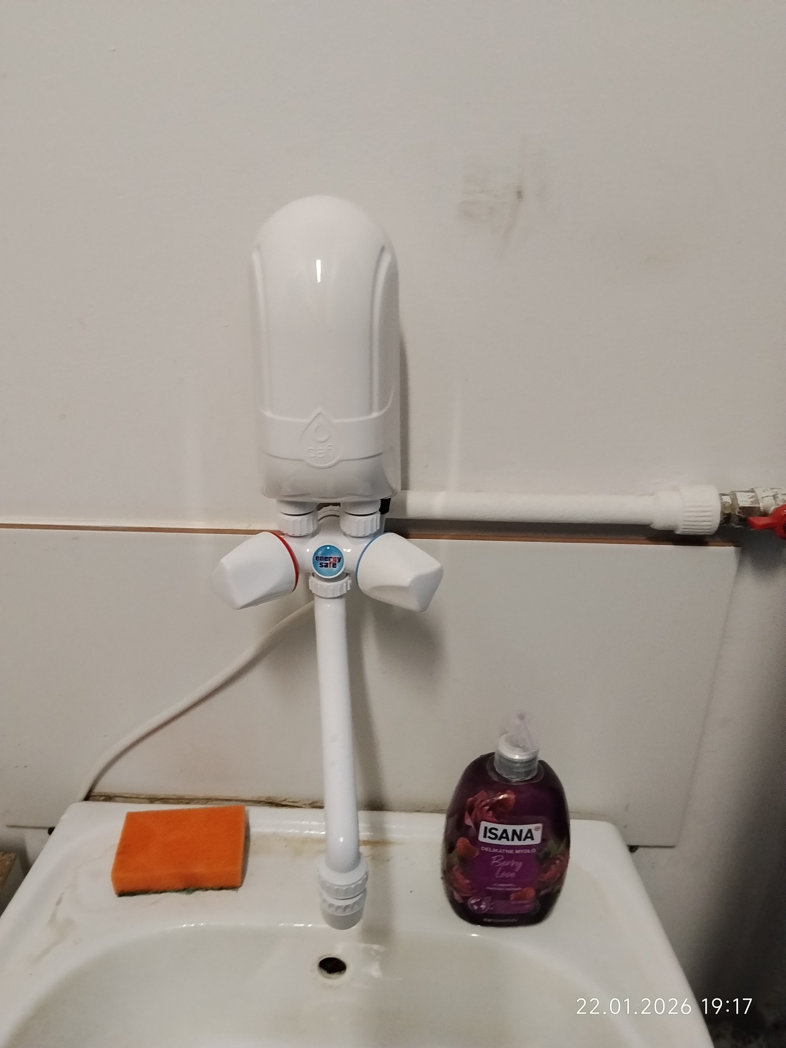 Biała bateria umywalkowa z podgrzewaczem wody, zamontowana na umywalce. Obok mydło w płynie i gąbka. Widoczna instalacja wodna.