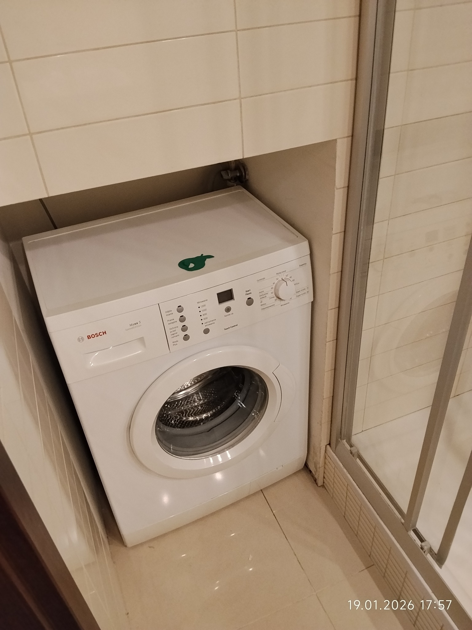 Biała pralka Bosch Maxx 5 wciśnięta w narożnik łazienki z beżowymi płytkami, widoczna kabina prysznicowa. Ujęcie z góry, lekko zniekształcona perspektywa.