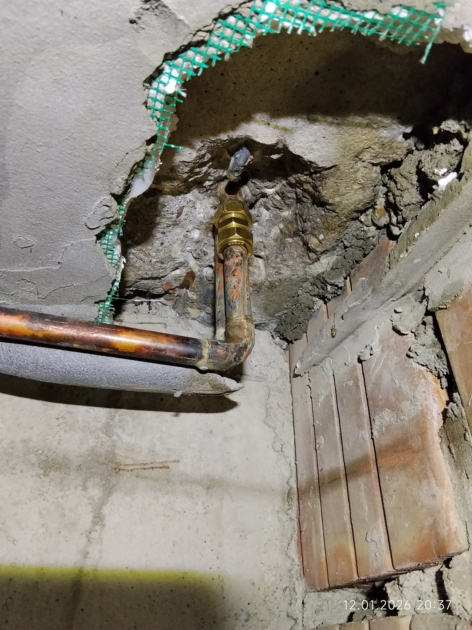 Miedziana rura z zaworem w ścianie z siatką w Warszawie. Widoczne ślady po kuciu i zaprawie. Instalacja po naprawie lub w trakcie przeróbki.