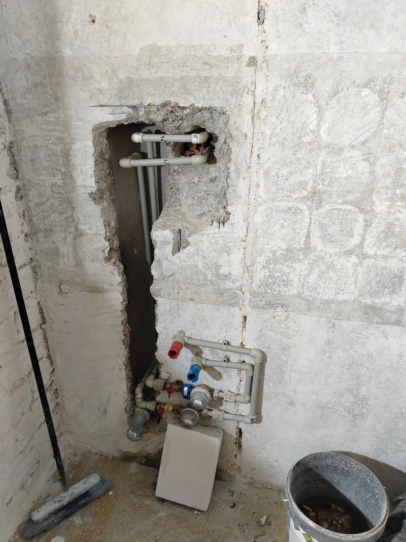 Szare rury instalacji wodnej wystają ze ściany w surowym stanie, z widocznymi elementami montażowymi i fragmentami gruzu. U dołu widoczny fragment wiadra i narzędzi.