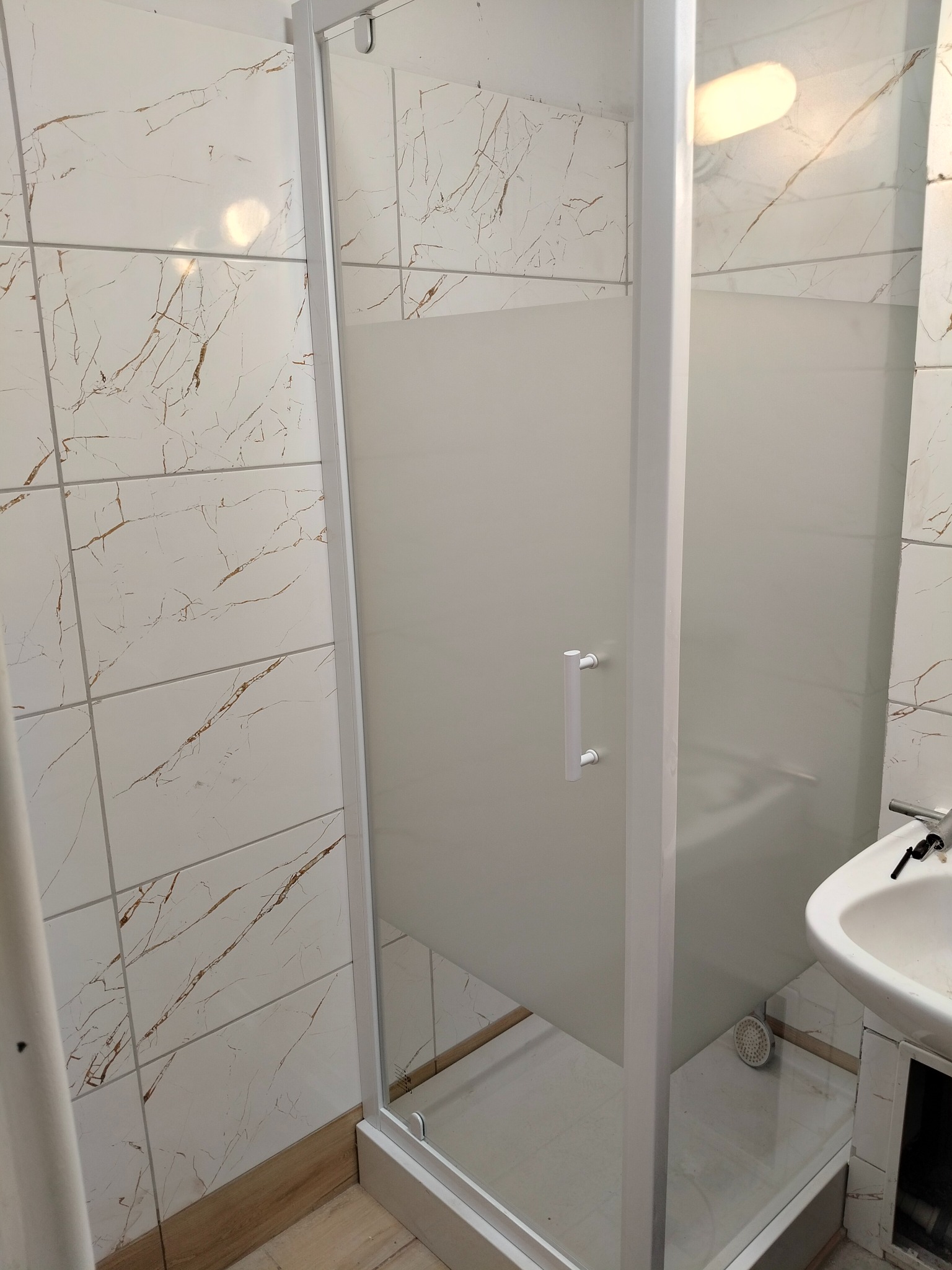 Nowoczesna kabina prysznicowa z matowego szkła i białymi profilami, umywalka z baterią w tle, płytki imitujące marmur na ścianach. Widok na łazienkę po remoncie.