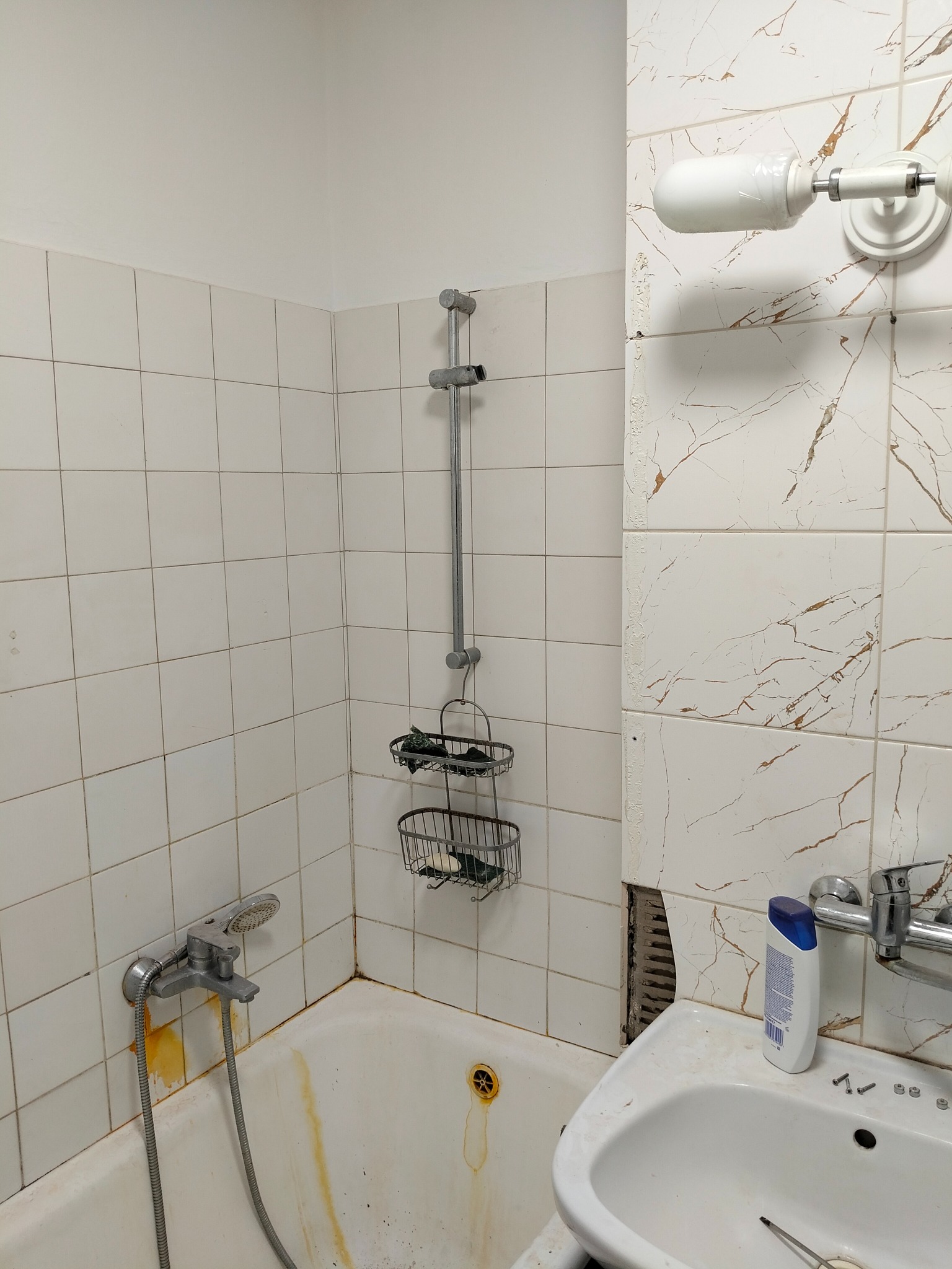 Zardzewiała bateria wannowa i słuchawka prysznicowa na tle białych, kwadratowych płytek. Widoczne ślady rdzy i niedoskonałości. Umywalka z brakującymi płytkami w tle.