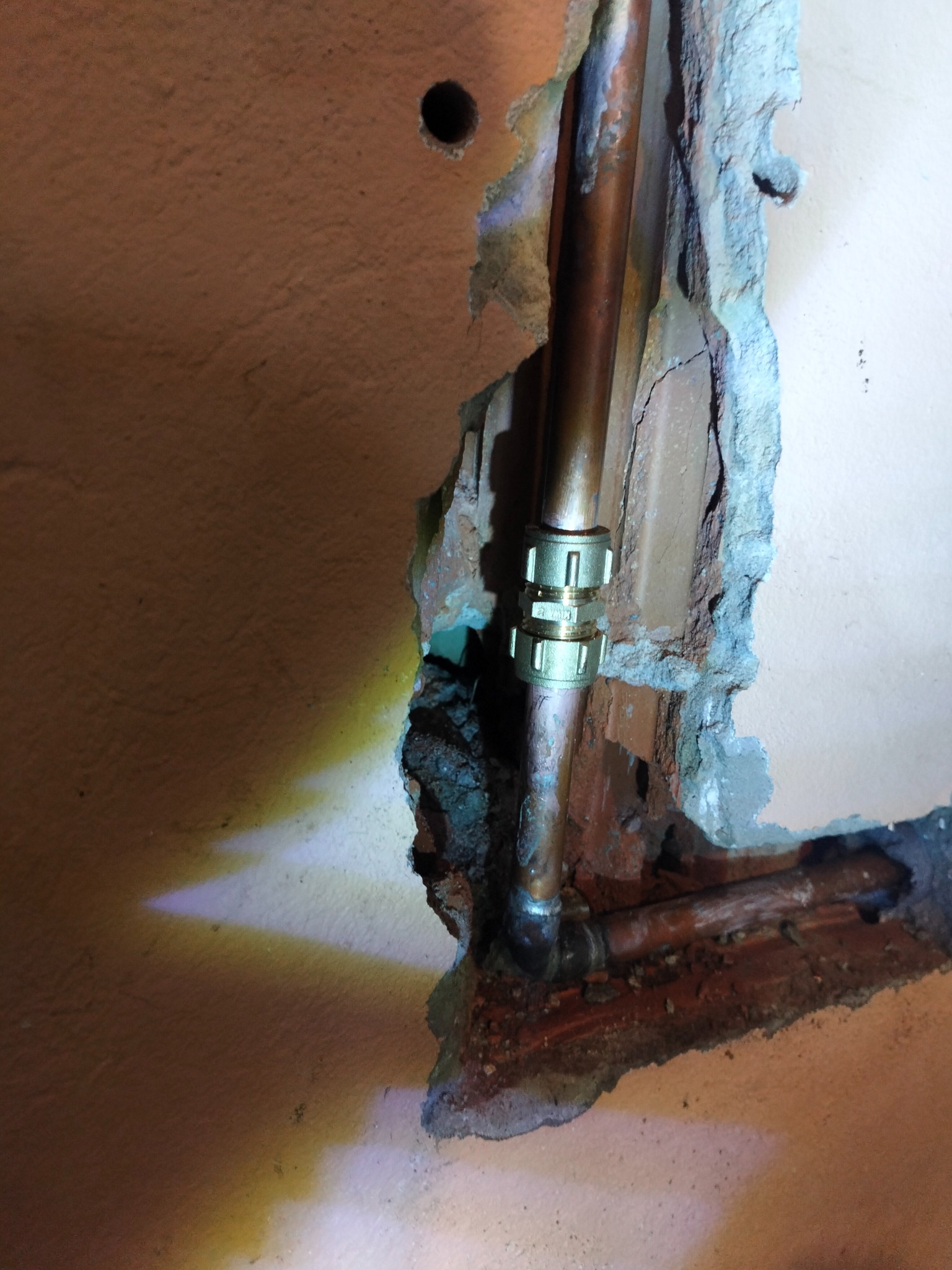 Miedziane rury połączone złączką w wykuciu ściany. Widoczne ślady po naprawie instalacji. Ujęcie z bliska, oświetlenie punktowe.