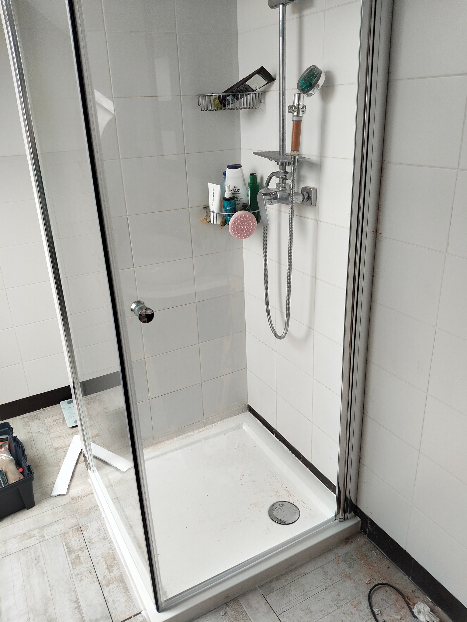 Kabina prysznicowa z chromowaną armaturą i półkami na kosmetyki na tle białych płytek. Widoczny brodzik i narzędzia hydrauliczne.