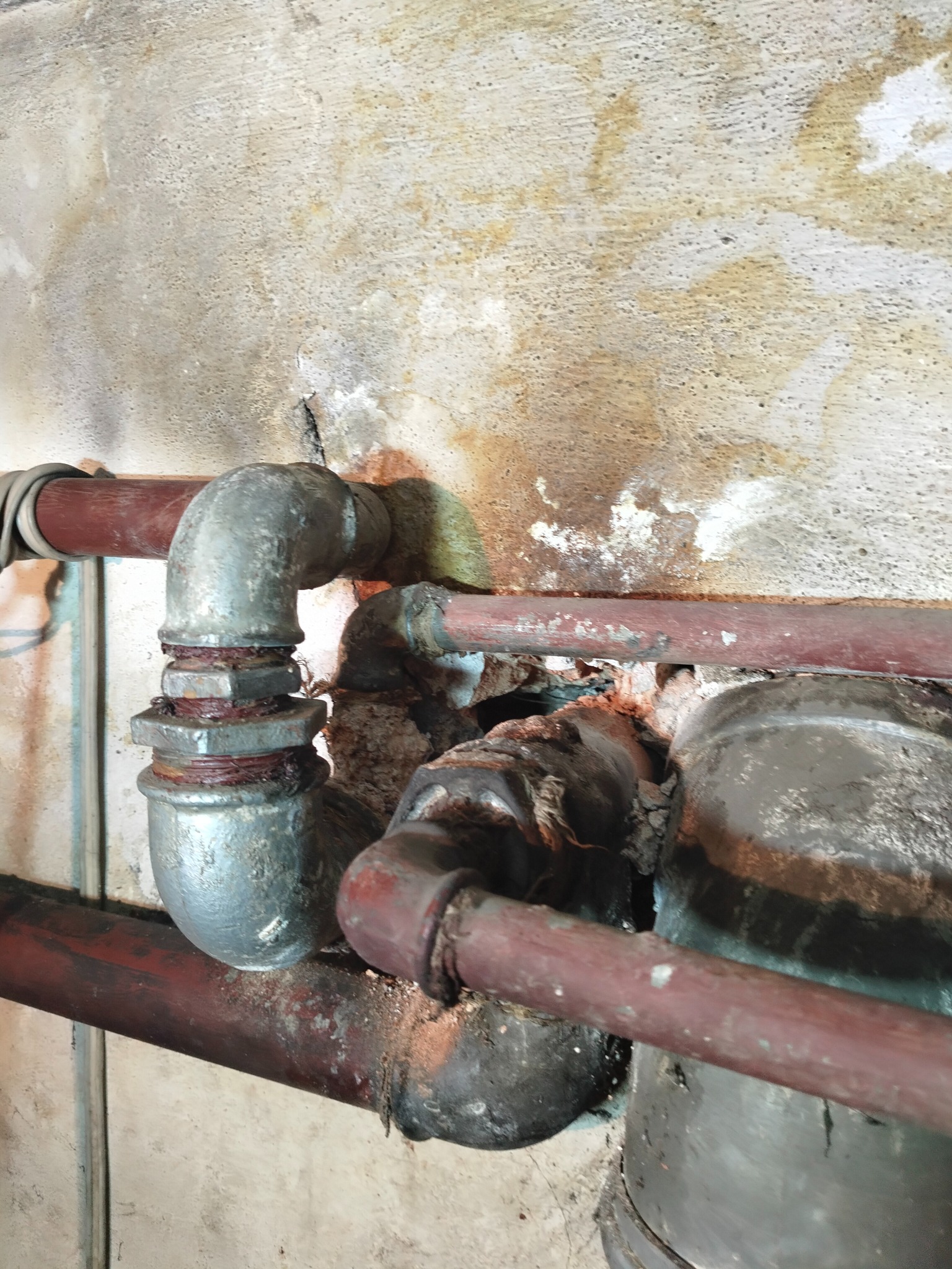 Stara, skorodowana instalacja grzewcza z rurami w kolorze czerwono-brązowym i szarymi złączkami, widoczna na tle ściany z zaciekami i pęknięciami.