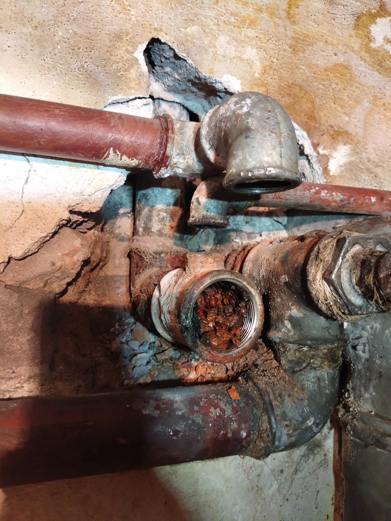 Zardzewiałe, metalowe rury hydrauliczne z otwartym gwintem, wypełnionym zanieczyszczeniami, osadzone w starej, popękanej ścianie. Widoczne ślady korozji i nieszczelności.