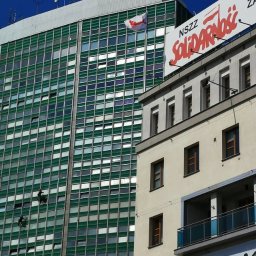 MIVLO Paweł Michalski - Dwaj alpiniści myją okna na wysokościowcu o zielonej fasadzie. W tle budynek z napisem Solidarność i masztami antenowymi na dachu, błękitne niebo.