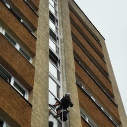 MIVLO Paweł Michalski - Pracownik na linie, w kasku, myje okna na wysokości w bloku z brązową elewacją. Widok z dołu, pochmurne niebo. Otwarta okna na różnych piętrach.