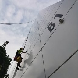 MIVLO Paweł Michalski - Pracownik w uprzęży czyści elewację budynku z napisem 'B1', używając gąbki. Widok z dołu, pochmurne niebo w tle.