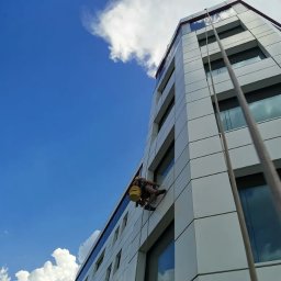 MIVLO Paweł Michalski - Pracownik na linach, z żółtym wiadrem, myje okna na wysokościowcu o nowoczesnej architekturze, na tle błękitnego nieba z chmurami.