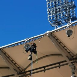 MIVLO - Pracownik na wysokości w Warszawie, zabezpieczony linami, wykonuje prace na konstrukcji dachu stadionu, widoczne oświetlenie. Prace wysokościowe.