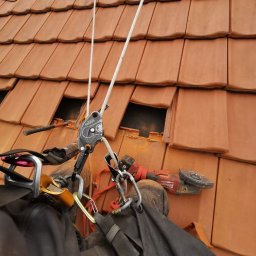 MIVLO - Praca na dachu z użyciem sprzętu alpinistycznego i szlifierki kątowej. Widoczne dachówki, liny, karabinki i narzędzie pracy. Bezpieczeństwo i precyzja podczas naprawy dachu.