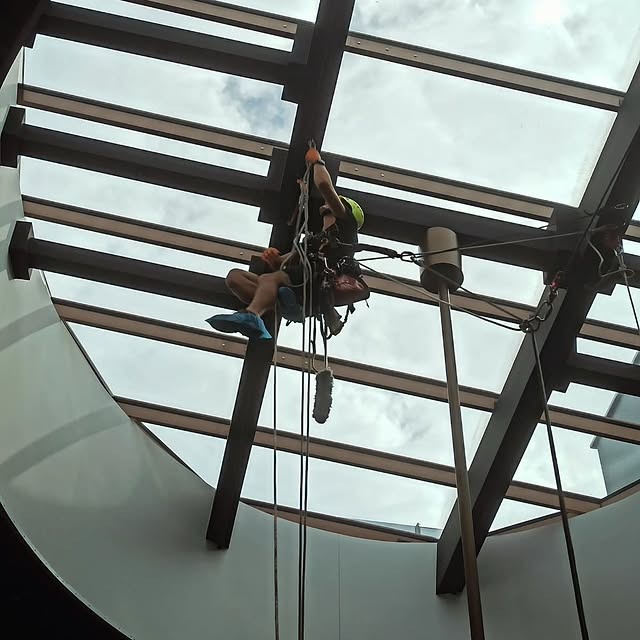 Pracownik na linach, w kasku, czyści szklany dach wewnątrz budynku. Widoczne liny, sprzęt alpinistyczny i elementy konstrukcji dachu.