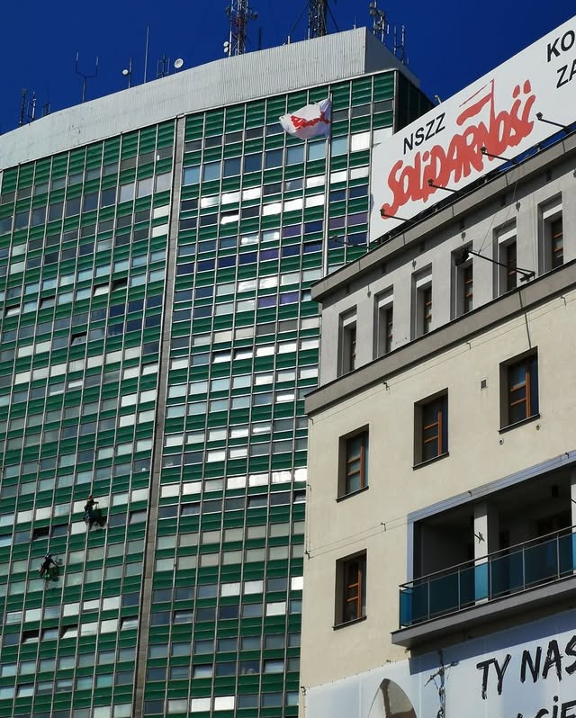 Dwaj alpiniści myją okna na wysokościowcu o zielonej fasadzie. W tle budynek z napisem Solidarność i masztami antenowymi na dachu, błękitne niebo.
