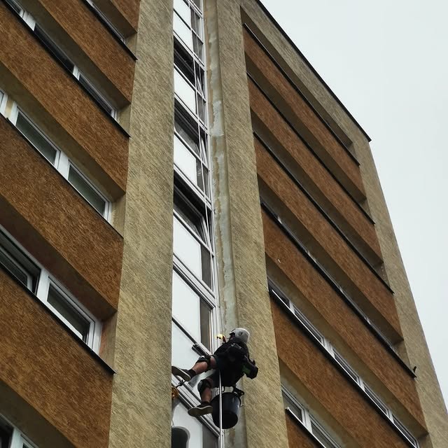 Pracownik na linie, w kasku, myje okna na wysokości w bloku z brązową elewacją. Widok z dołu, pochmurne niebo. Otwarta okna na różnych piętrach.